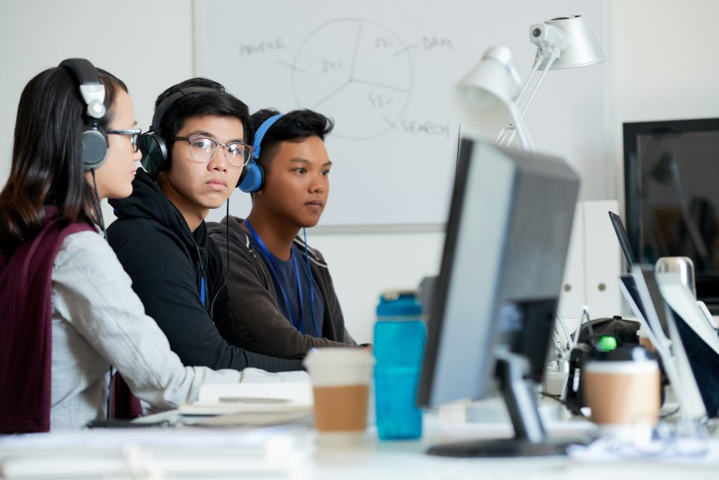 Three young Asian software developers wearing headphones work together at computer desks in a modern office. A whiteboard with a system diagram is in the background.